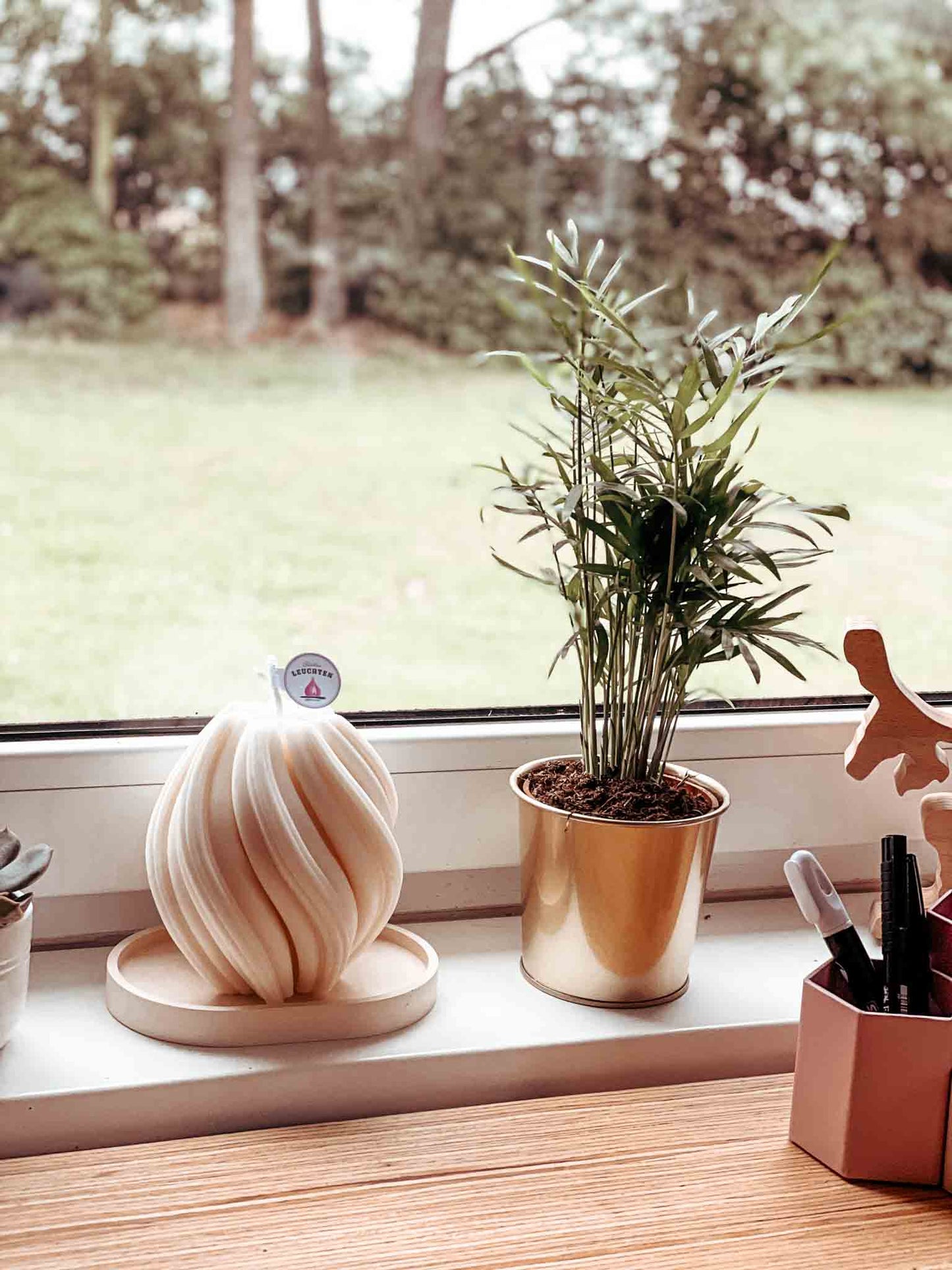 Wellenkerze in cremeweiß neben Topfpflanze auf dem Fensterbrett im Hintergrund Rasen und Bäume