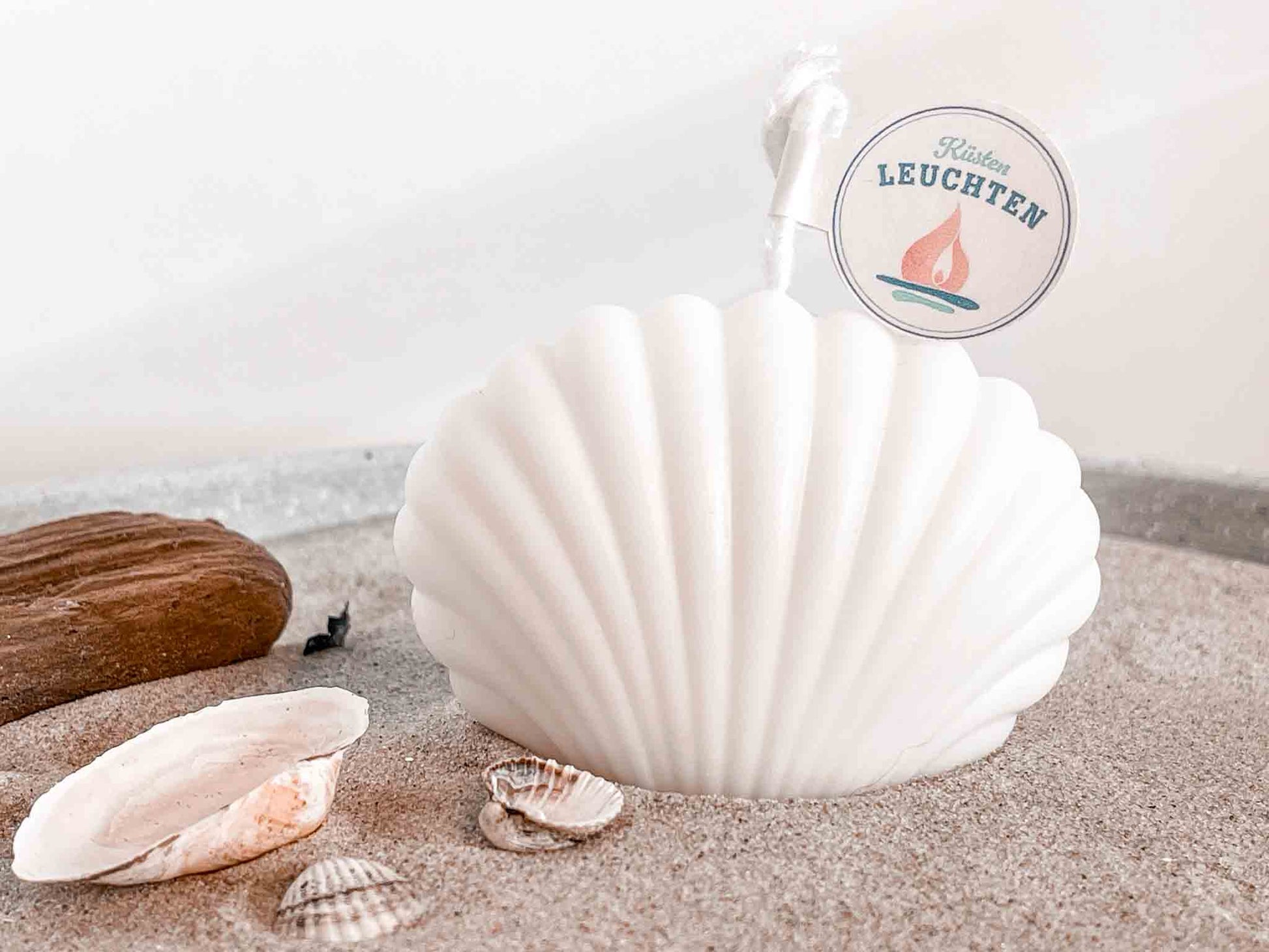 kleine Muschelkerze in cremeweiß auf Strandsand Strandgut im linken VOrdergrund