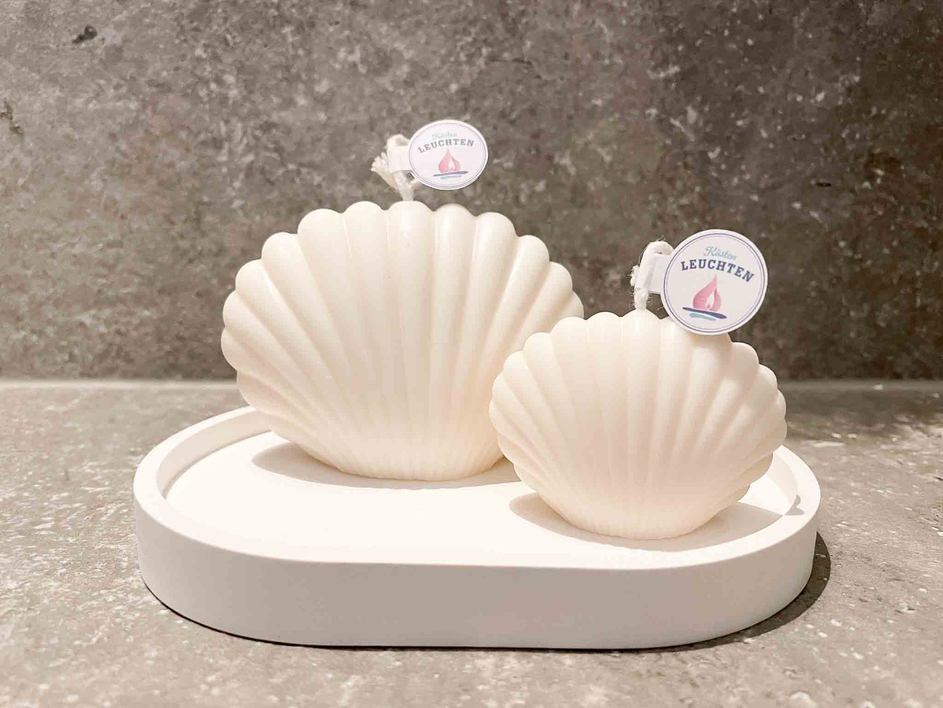 2 Muschelkerzehn in cremeweiß auf weißem ovalem Teller dunkelbraune Fliesen im Hintergrund