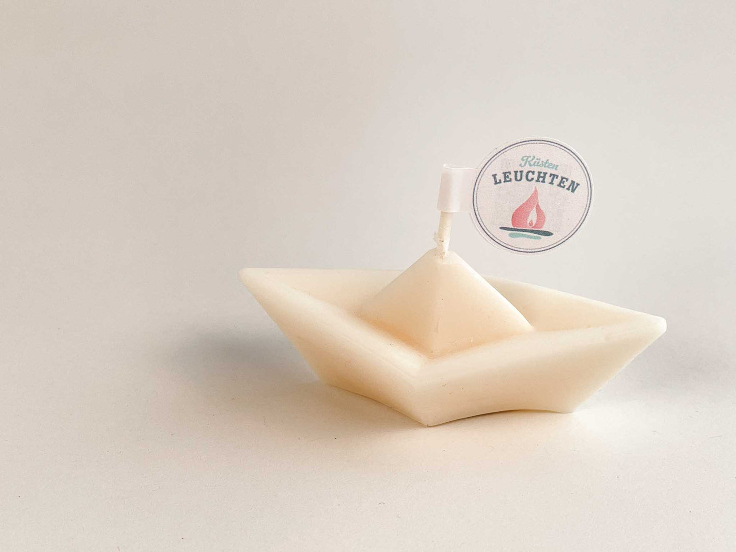 cremeweißes Schiffchen aus Rapswachs mit KuestenLeuchten Label vor weißem Hintergrund