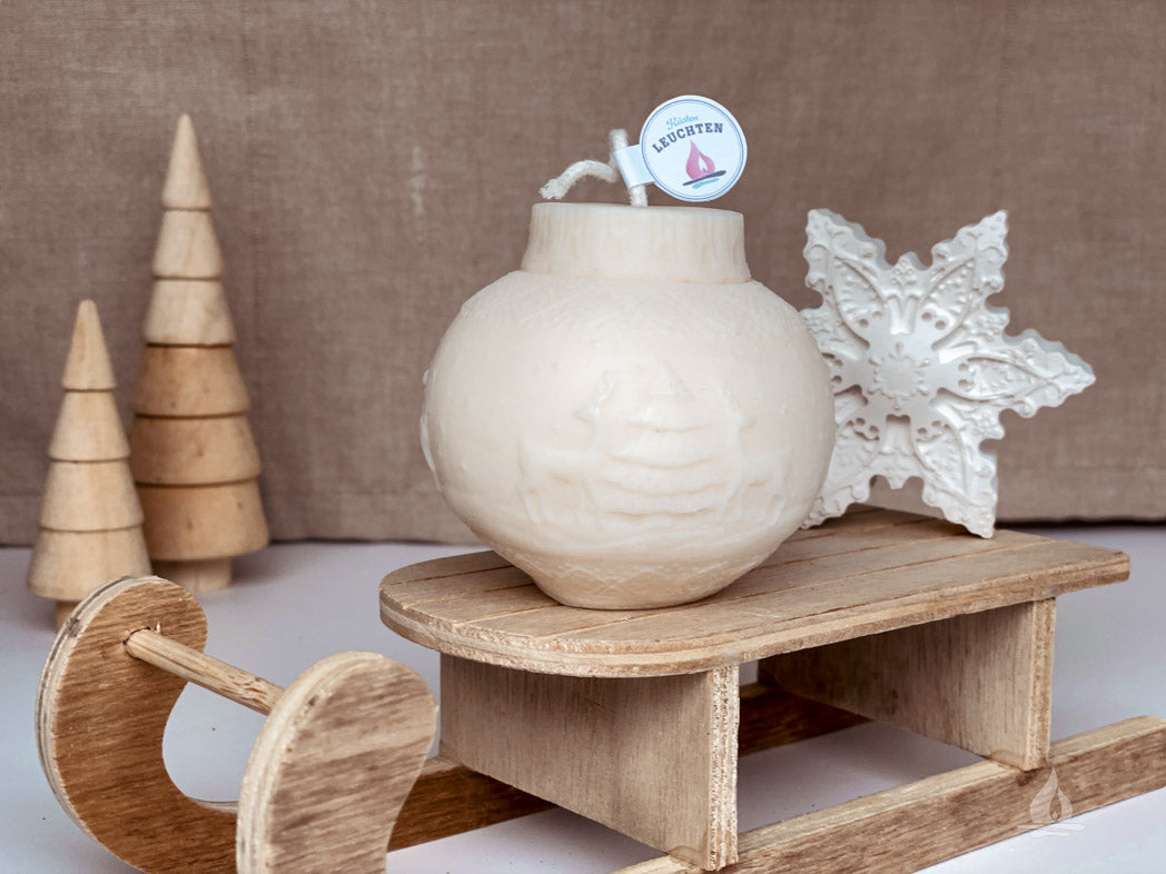 Kugelkerze mit Rentiermotiv auf Miniaturschlitten Tannenbäume aus Holz im Hintergrund