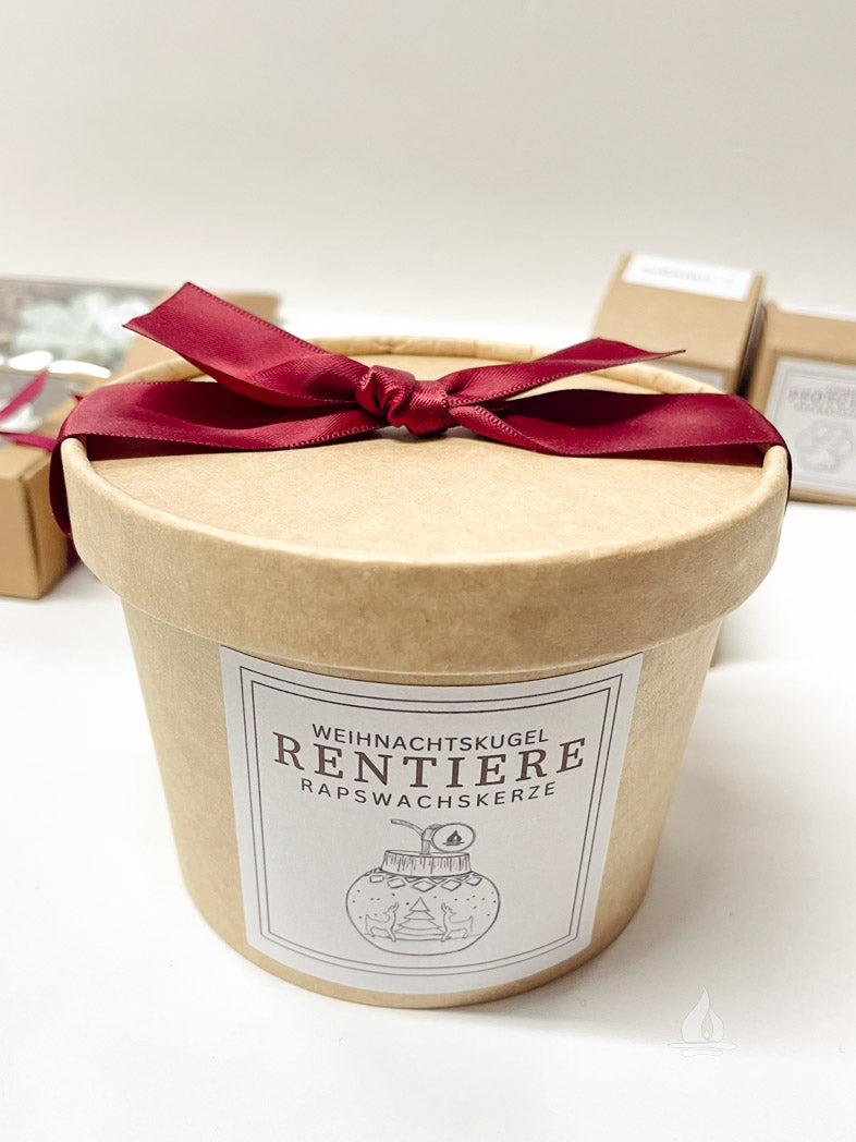 runde Verpackung aus Kraftpapier mit roter Schleife auf dem Deckel und schwarz-weißem Label mit gezeichnetem BIld von der Kugelkerze mit Rentiermotiv