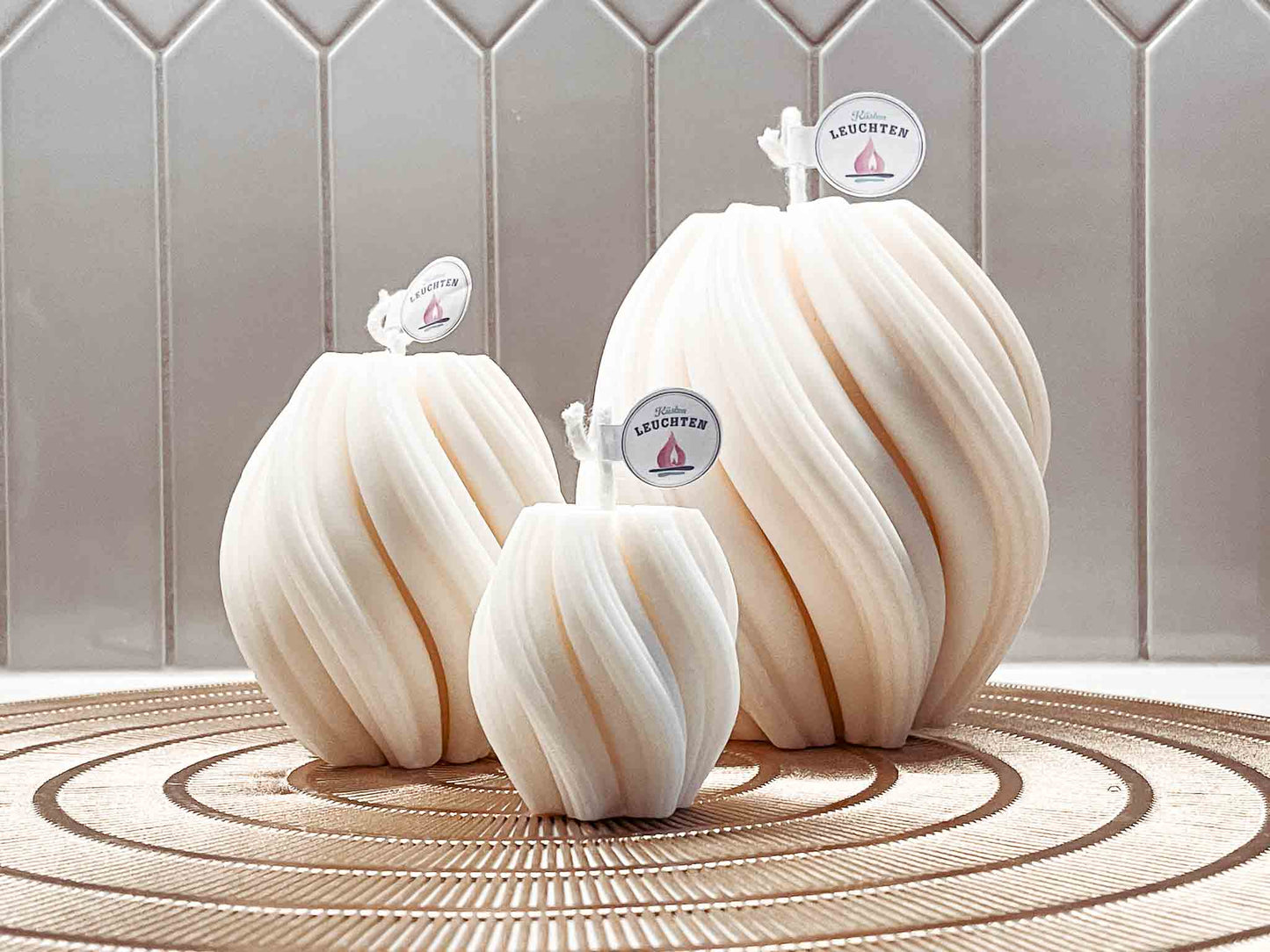 3 Wellenkerze in unterscheidelichen Größen vor grauen Fliesen auf runder goldfarbener Unterlage