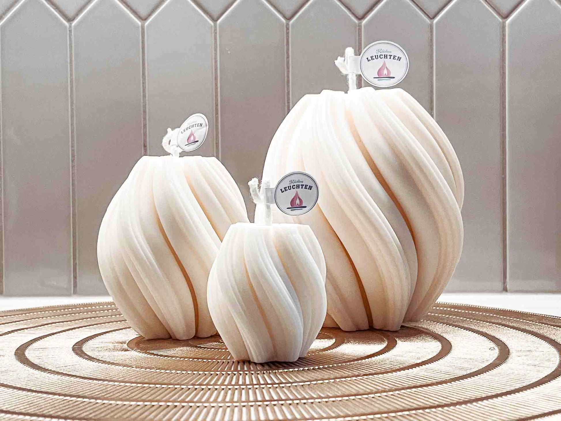 3 Wellenkerze in unterscheidelichen Größen vor grauen Fliesen auf runder goldfarbener Unterlage