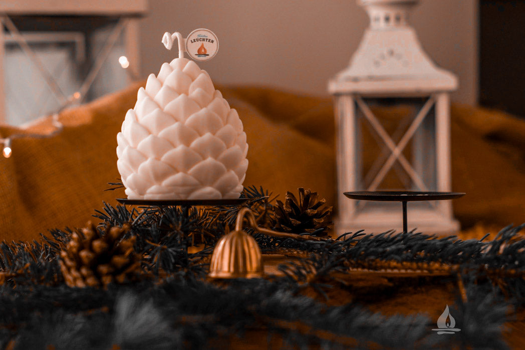 Rapswachskerze als Zannenzapfen mit weihnachtlichem Grün und weißer Laterne im Hintergrund