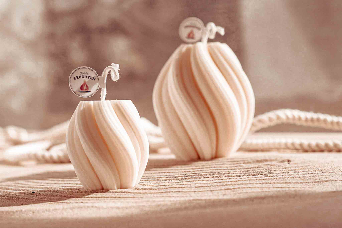 zwei Kerzen in gedrehter Form in zwei Größen auf Sand im Hintergrund weißes Seil