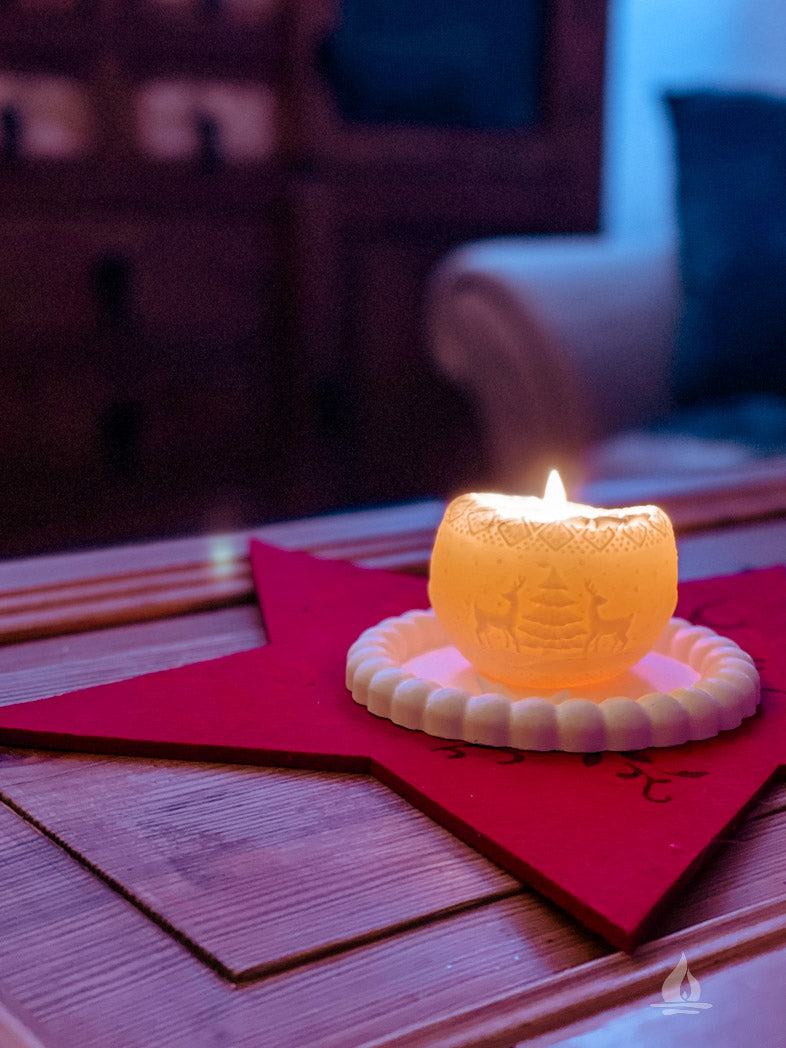 cremeweiße Kugelkerze mit Rentiermotiv brennend auf rotem Sternuntersetzer auf Holztisch